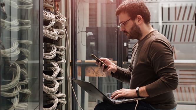 IT professional kneeling next to server rack, reviewing diagnostics on his phone and laptop